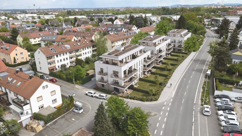 Luftaufnahme einer Wohnsiedlung mit Gärten, weißen Gebäuden, umgeben von Straßen und grünen Bäumen.